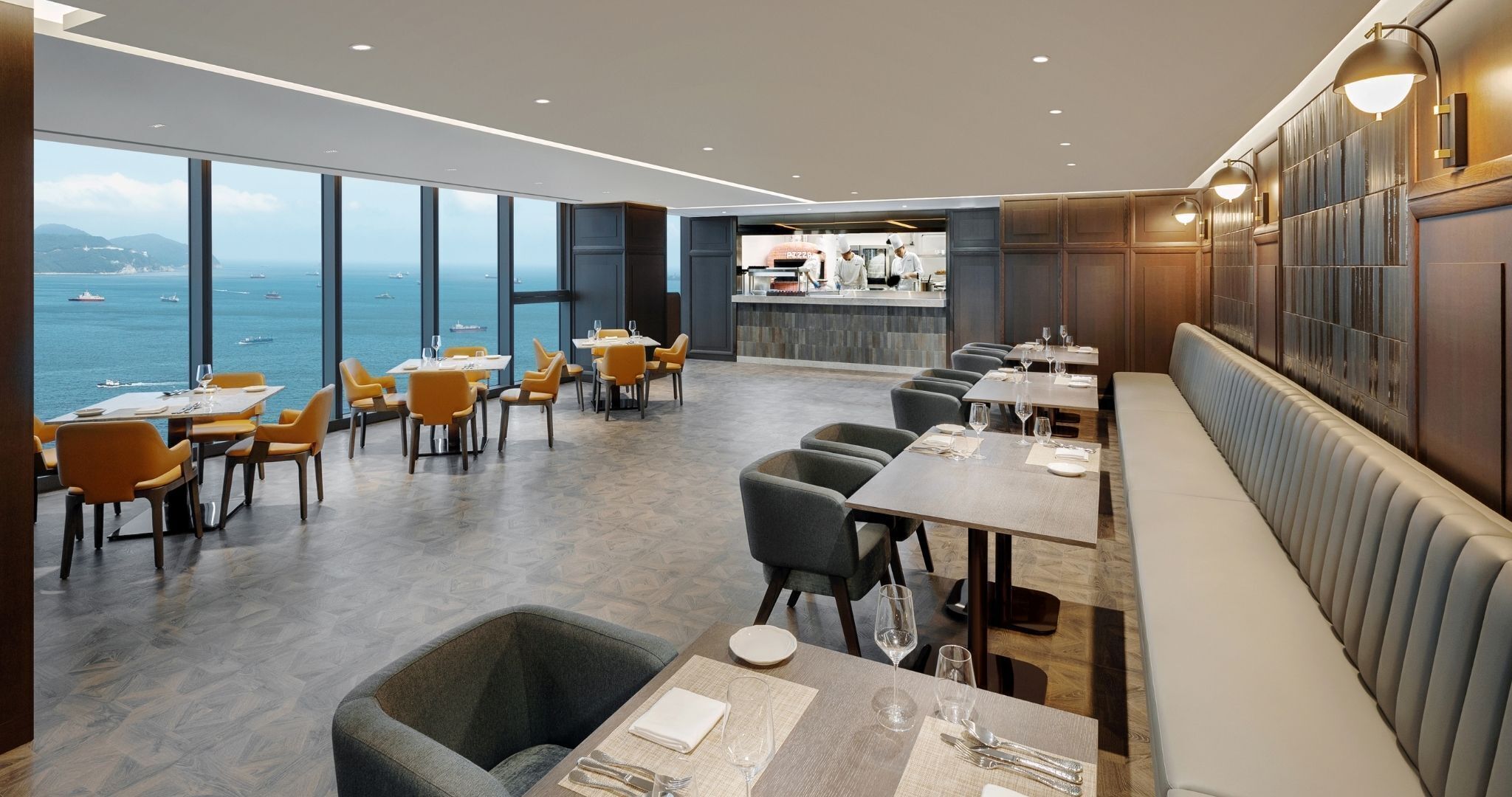 A modern restaurant with a view of the sea, featuring a mix of dining tables and booths, and a bar area in the background.