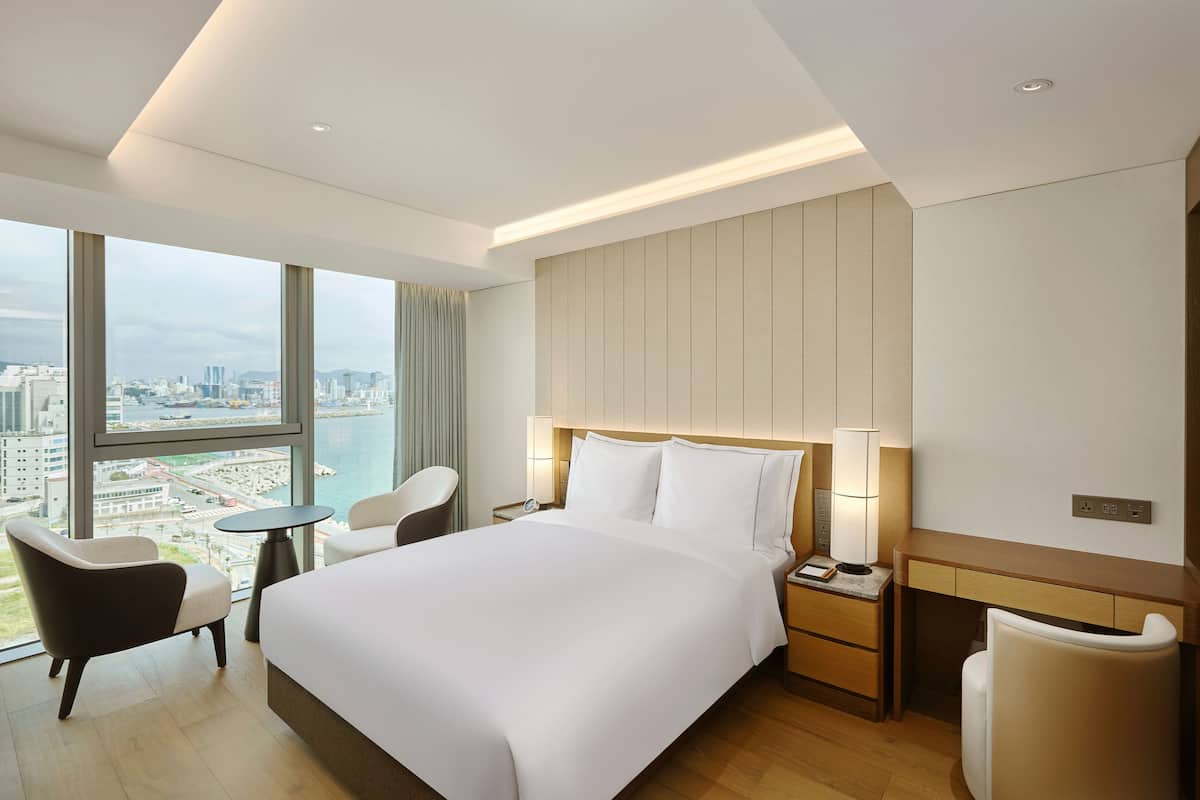 Ein modernes Hotelzimmer mit einem großen Bett, einem Schreibtisch, einem Stuhl und Blick auf die Stadt durch große Fenster.