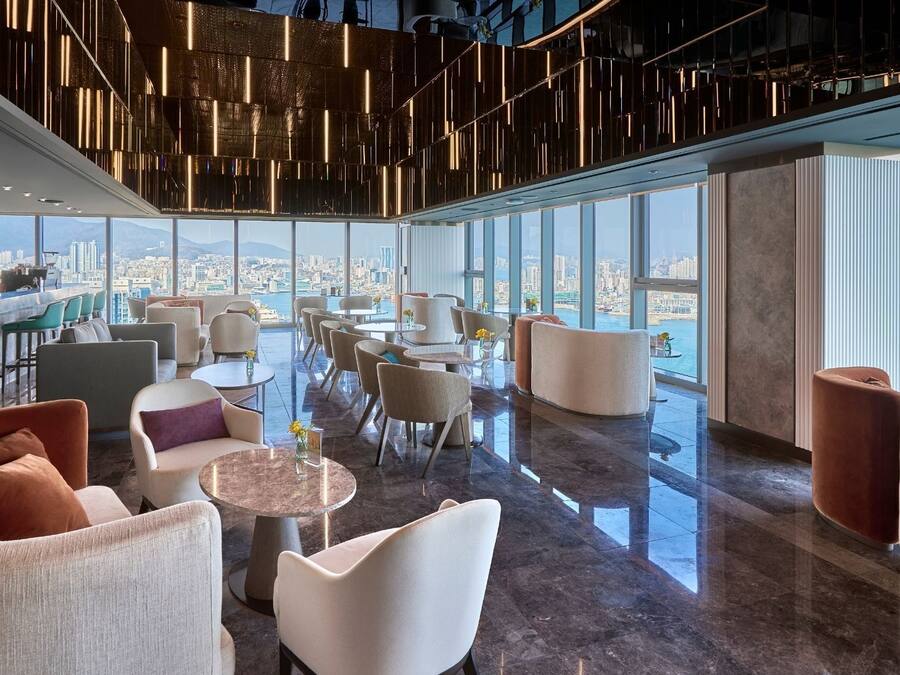 Un salón moderno con vista a la ciudad, que cuenta con una zona de bar, arreglos de asientos y un techo alto con luces colgantes.