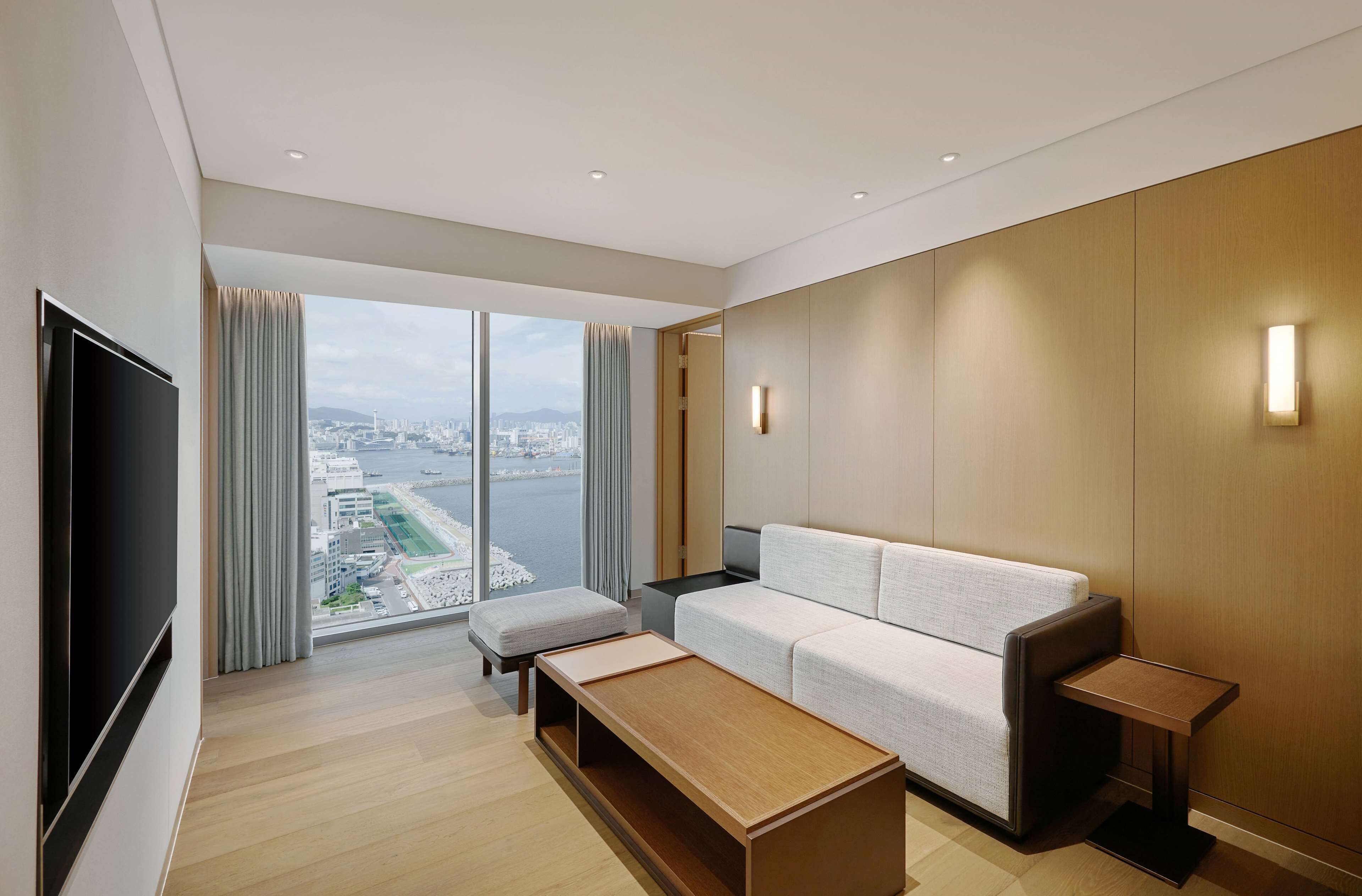 A modern hotel room with a large window offering a city view, a flat-screen TV, a sofa, a coffee table, and a small side table.