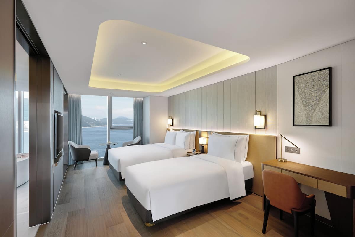 Ein Hotelzimmer mit zwei Betten, einem Schreibtisch, einem Stuhl, einem Fernseher und Meerblick.