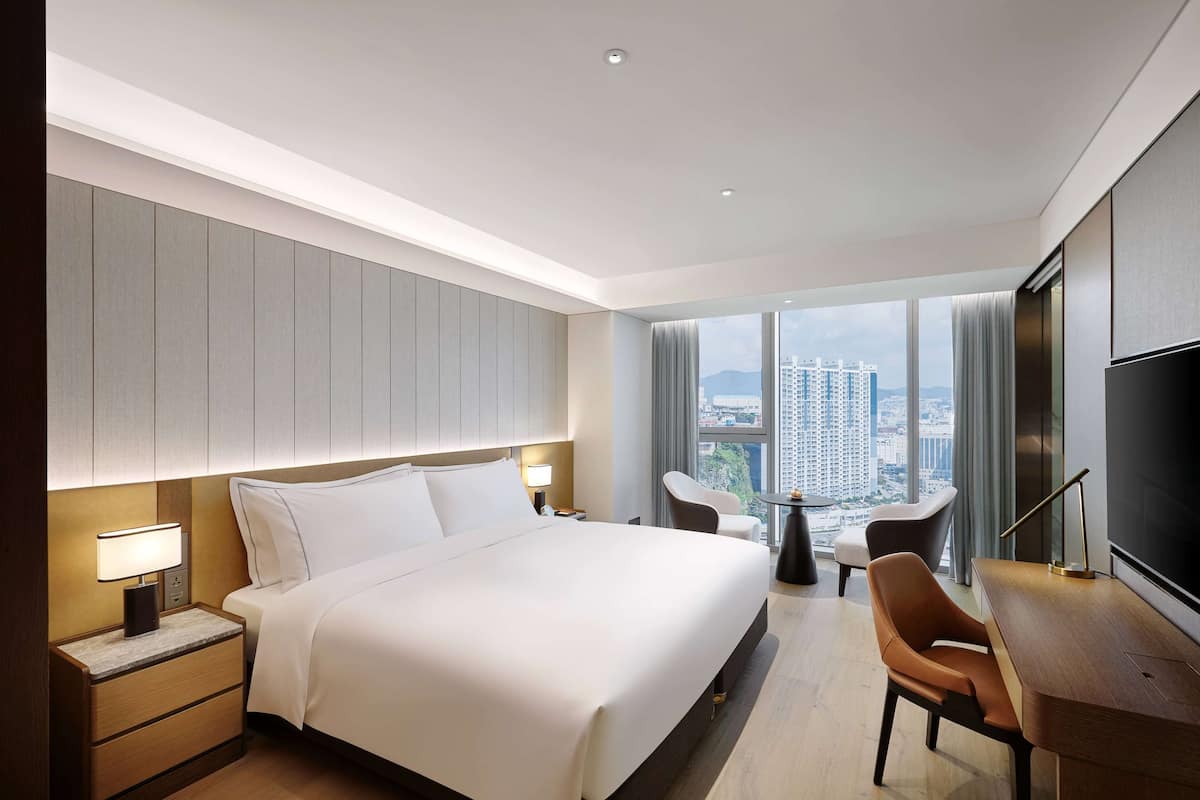 Ein modernes Hotelzimmer mit einem großen Bett, Nachttischen, einem Schreibtisch mit Stuhl, einem Fernseher und Blick auf die Stadt durch große Fenster.