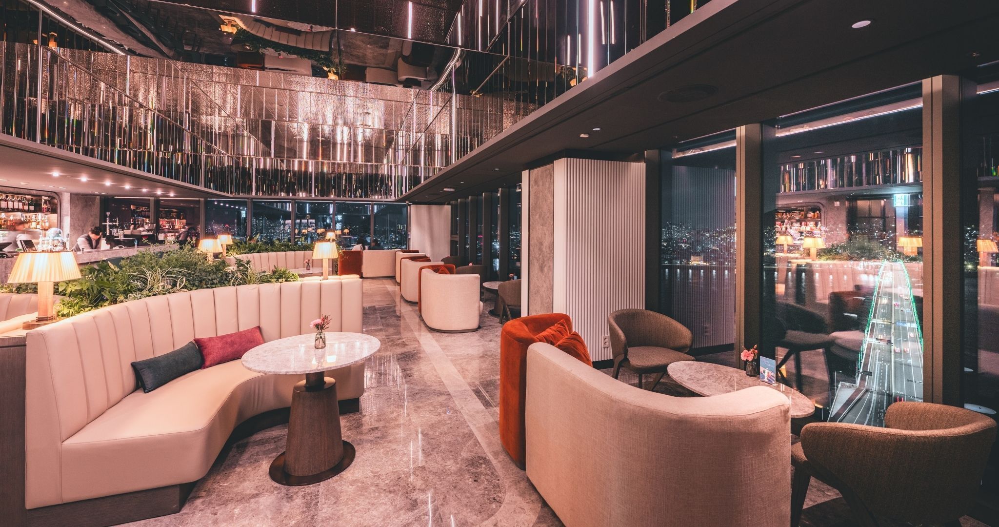 A modern lounge area with seating and a bar, featuring a city view through large windows.