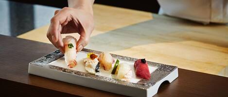 A hand holding a sushi piece with a garnish, placed on a rectangular plate with other sushi pieces and garnishes.