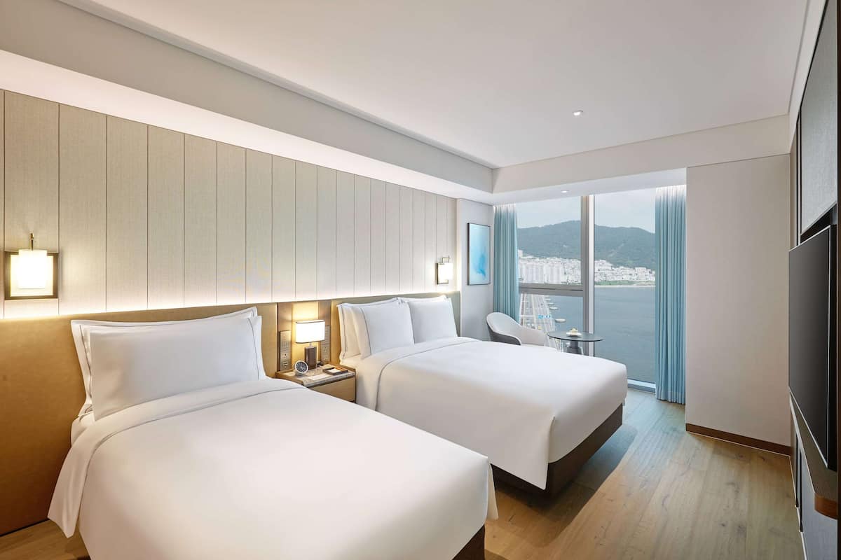 Ein Hotelzimmer mit zwei Betten, einem Fernseher, einem Schreibtisch mit Lampe und Blick auf eine Stadtlandschaft durch große Fenster.