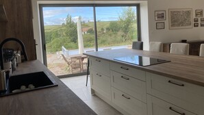 Une cuisine moderne dotée d’un grand îlot central, d’un coin repas avec une table et des chaises, et offrant une vue sur un paysage verdoyant grâce à de grandes fenêtres.