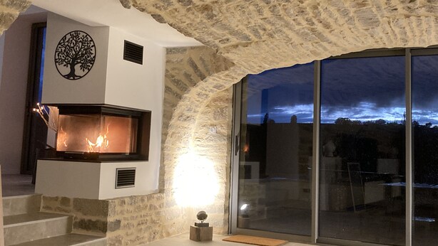 Une pièce aux murs de pierre, dotée d’une cheminée, d’un motif décoratif représentant un arbre et offrant une vue sur le ciel par le biais d’une grande fenêtre.