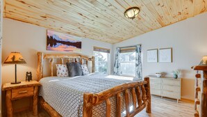Una cama con estructura de madera y colcha estampada, una mesita de noche con lámpara, un aparador y un cuadro de un paisaje montañoso.