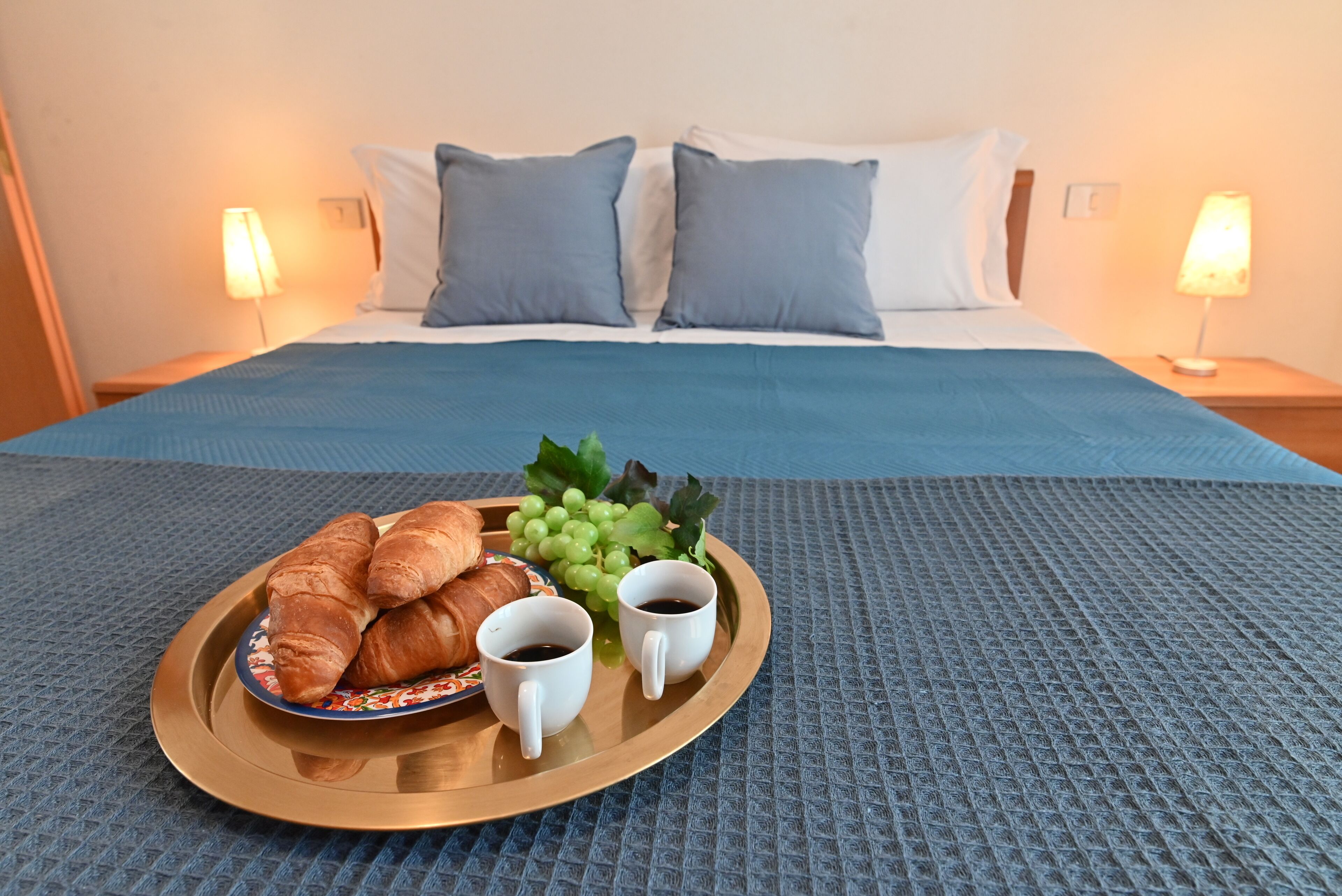 Un letto rifatto con una coperta blu, due cuscini e un vassoio con croissant, uva e due tazze di caffè su un tappetino blu con texture.