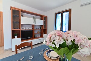 Un comedor con una mesa lista para dos personas, un televisor al fondo y una estantería con los mensajes "IZZ HOME" y "LOVE".