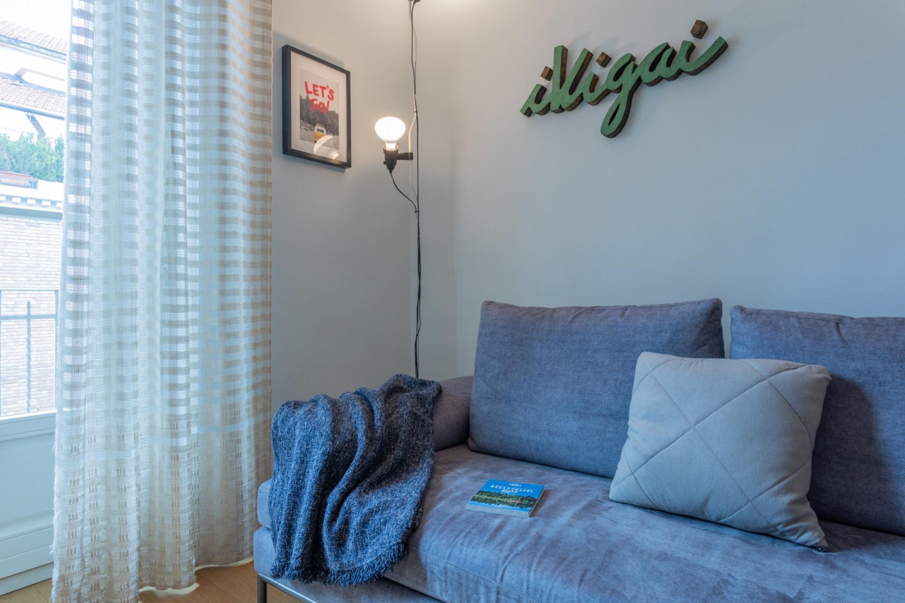 Una habitación con un sofá gris, una manta azul, un cuadro enmarcado en la pared y una lámpara fijada a la pared.