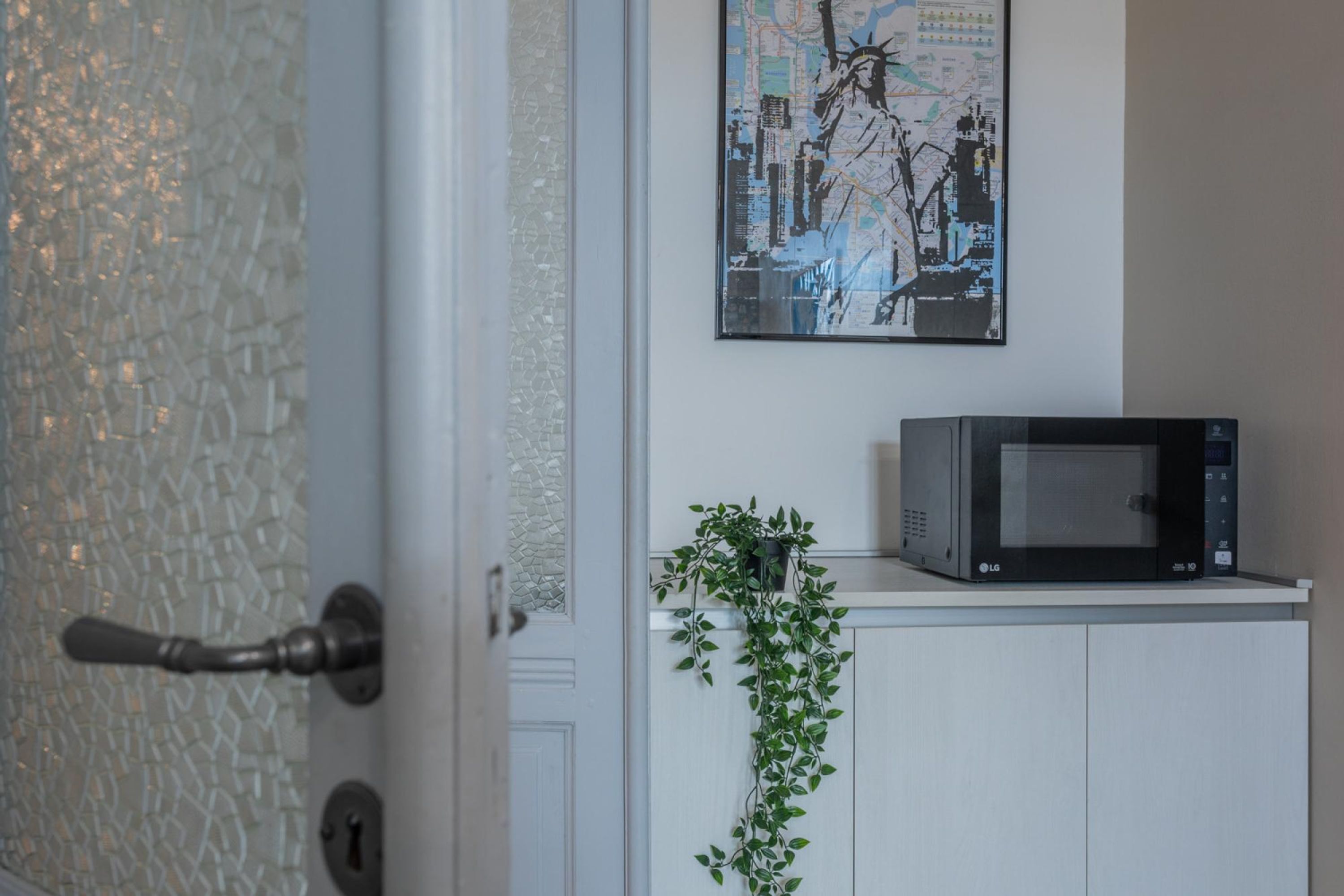 Une porte d’entrée avec une vitre, un meuble blanc contenant un four à micro-ondes, et un tableau accroché au mur.