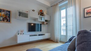 A modern living room with a grey sofa, a flat-screen TV on a white entertainment unit, and a large window with curtains. - La Casa dei Viaggiatori by Wonderful Italy (Turin)