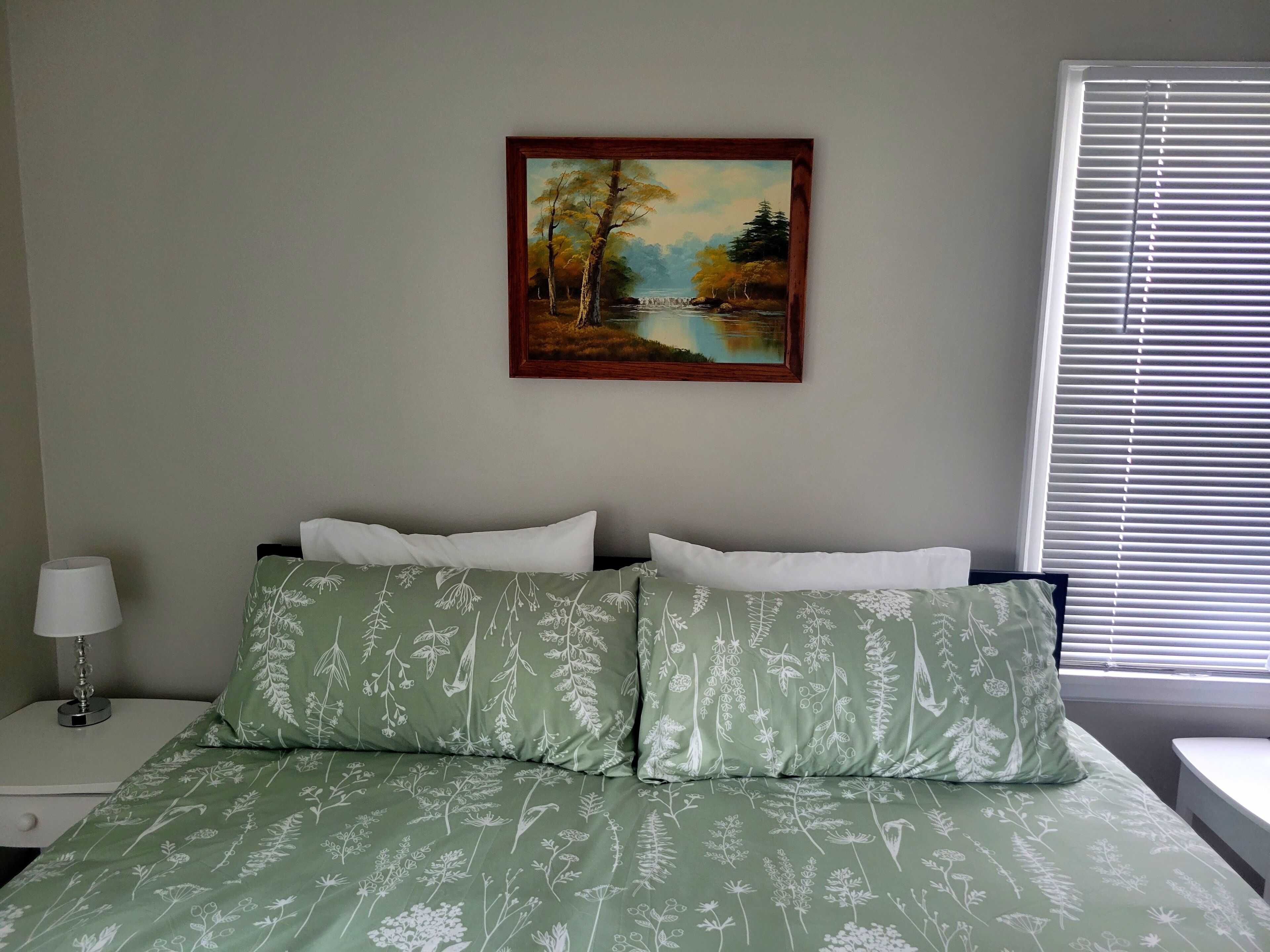 Un dormitorio con una cama, dos almohadas, una mesita de noche con una lámpara y una ventana con persianas.