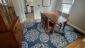 A dining area with a wooden table and chairs, a patterned rug, and a view into the kitchen.
