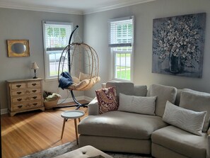 A living room with a hanging chair, a sofa, a small table, a dresser, and a painting on the wall. - Kutzky Cottage | 3 BR , Steps to Mayo St Marys (Rochester)