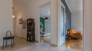 Un couloir avec une bibliothèque, une chaise et un canapé, dans une pièce dont le sol est carrelé et qui possède une porte donnant sur une autre pièce.