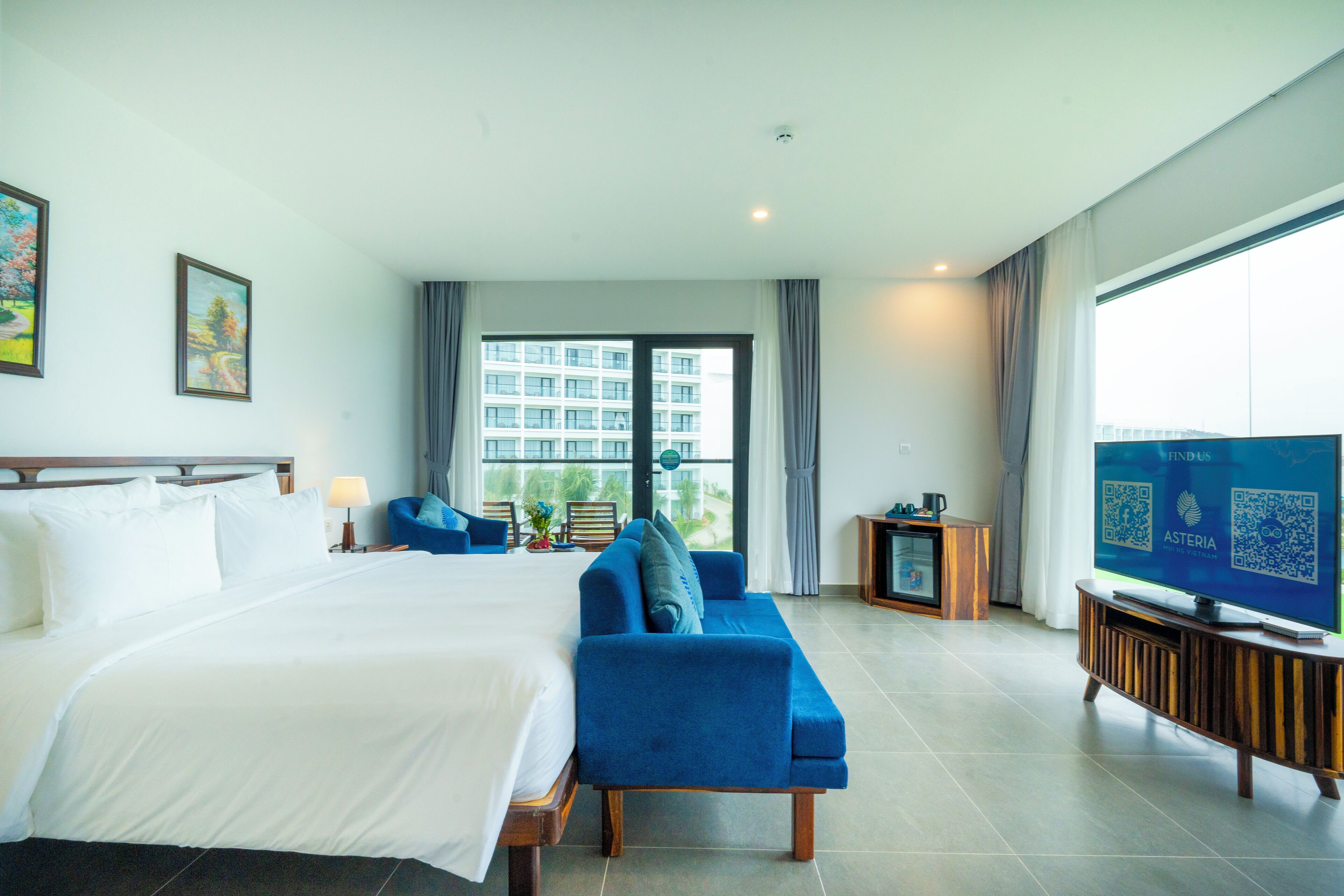 A hotel room with a large bed, a blue sofa, a television, and a view of a building through the window.