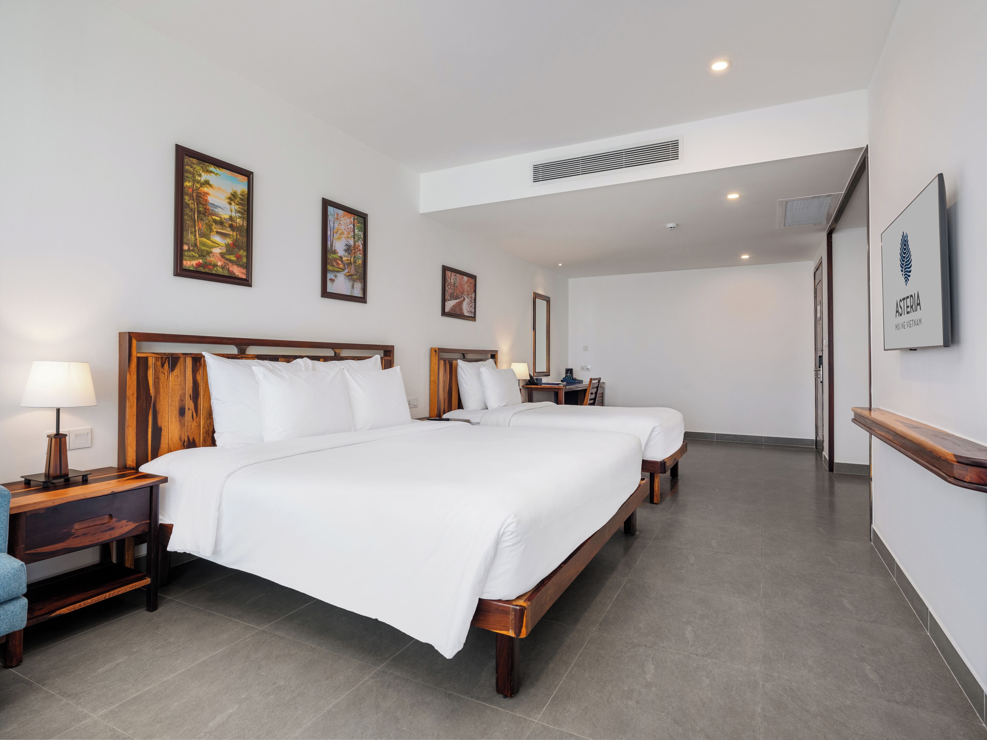 A hotel room with two beds, a desk, a chair, a nightstand, and a painting on the wall.