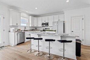 A modern kitchen with white cabinetry, a central island with bar stools, stainless steel appliances, and a window with blinds. - Hideaway Spot Where Southern Charm Stays -  LT7950 (Calcasieu Parish)