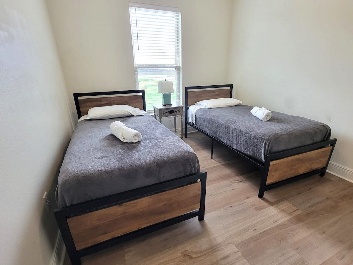 A room with two beds, a nightstand, and a window with blinds.