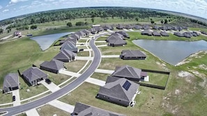 A residential area with houses arranged in a curved layout, connected by roads, and surrounded by green fields and bodies of water.