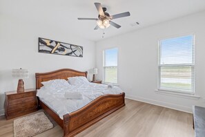 Ein Schlafzimmer mit einem hölzernen Bett, Deckenventilator, Nachttisch, Lampe, Teppich und zwei Fenstern mit Jalousien.