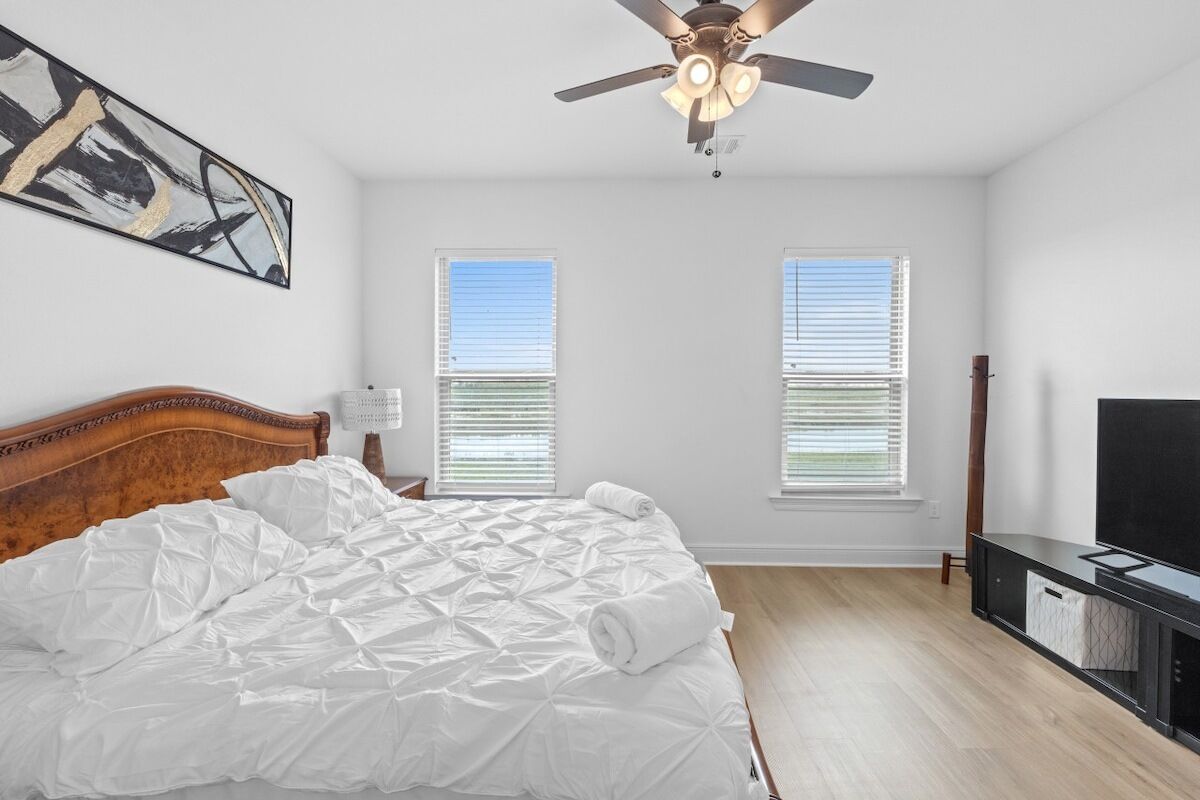 A bedroom with a bed, ceiling fan, two windows with blinds, and a TV on a stand.