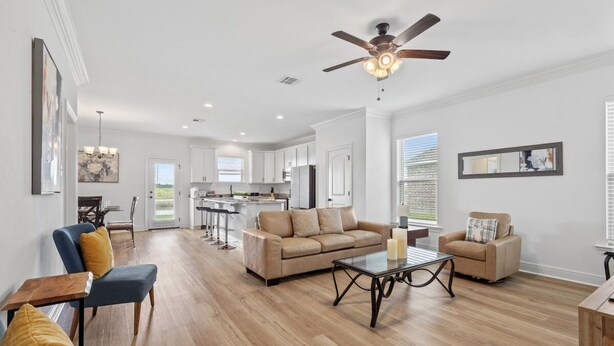 A spacious living room with a sofa, armchairs, and a coffee table. There is a ceiling fan and a kitchen in the background.