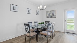 A dining area with a glass table surrounded by chairs, a chandelier, framed artwork on the walls, and a door with a view.