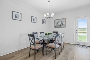 Ein Essbereich mit einem Glastisch, umgeben von Stühlen, einer Chandelierleuchtre und gerahmten Kunstwerken an den Wänden, sowie einer Tür mit Blick.