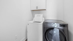 A laundry room with a front-loading washing machine and a dryer, a white cabinet above the dryer, and a power outlet on the wall. - Hideaway Spot Where Southern Charm Stays - LT7950 (Calcasieu Parish)