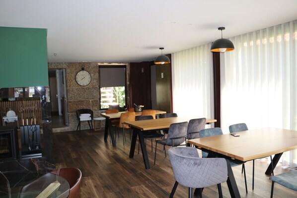 A modern dining area with wooden tables and chairs, a clock on the wall, and large windows with sheer curtains. - Duas Quintas-areal T1 Varanda (Nespereira GMR)