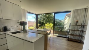A modern kitchen with a large island, a dining area with a table and chairs, and a balcony with a view of greenery and buildings. - Duas Quintas-areal T1 Varanda (Nespereira GMR)
