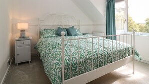 Ein Schlafzimmer mit weißem Metallbettgestell, einer Bettdecke mit grünem Blattmuster, einem Nachttisch mit Lampe und einem Fenster mit Vorhängen.