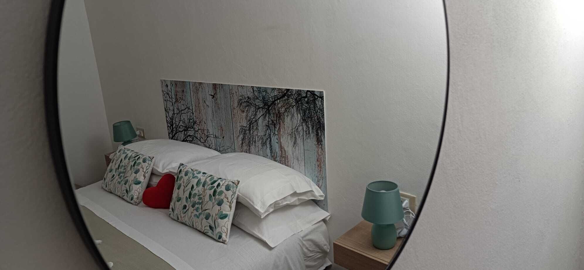 Uma cama bem arrumada com almofadas brancas e estampadas, uma almofada em formato de coração vermelho e um abajur azul-esverdeado sobre uma mesa de cabeceira.