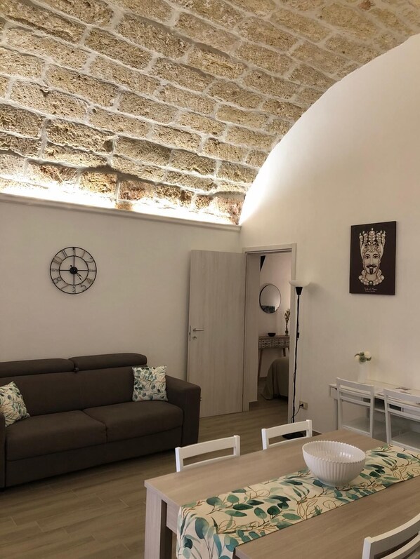 A cozy living room with a brick ceiling, a wooden dining table with chairs, a brown sofa with cushions, a wall clock, and a framed artwork. - Le Antiche Alcove - Charming detached house near the sea (Terrasini)