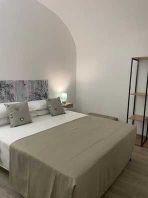 A modern bedroom with a large bed, a nightstand with a lamp, and a wooden shelf unit. - Le Antiche Alcove - Charming detached house near the sea (Terrasini)