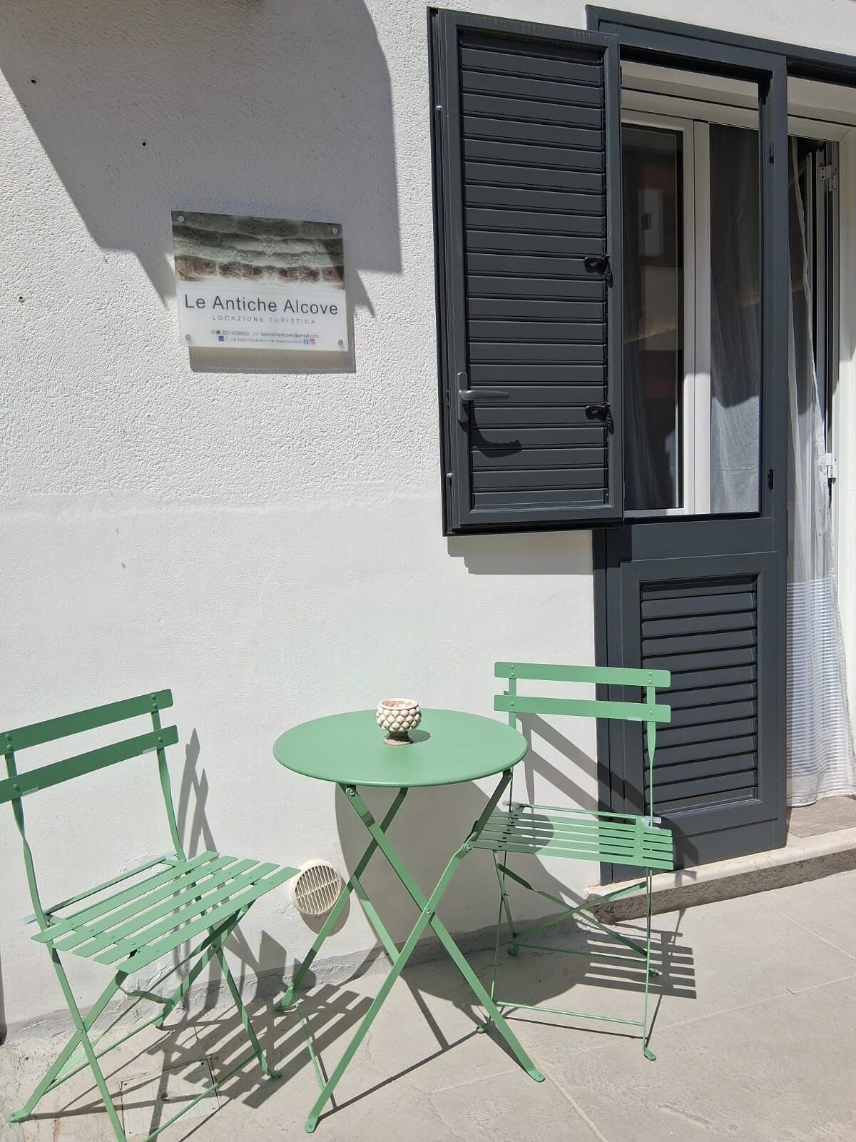Een klein buitgedeelte met twee groene metalen stoelen en een ronde tafel, een deur met zwarte luiken en een bord aan de muur.