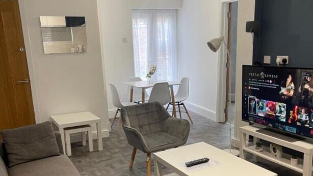 Een woonkamer met een grijze bank, een witte salontafel en een tv waarop een film draait. Op de achtergrond is een eethoek met een tafel en stoelen te zien.