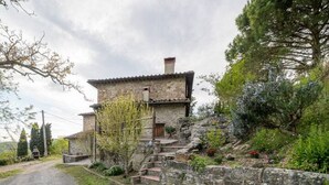 Una casa de piedra rústica con techo inclinado y chimenea, rodeada de abundante vegetación y un camino de grava.