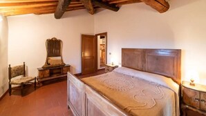Un dormitorio con techo de madera, una cama con cabecera, una silla, una mesita de noche y una puerta que da a otra habitación.