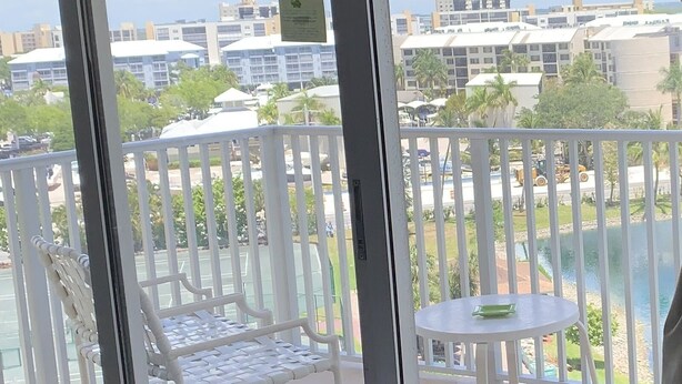 A balcony with a white chair and table, a patterned pillow on a sofa, and a view of buildings and greenery. - Beachfront Condo in Fort Myers Beach (Fort Myers Beach)