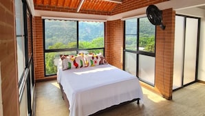 A bedroom with a large bed, a fan, and a view of a forest through large windows.