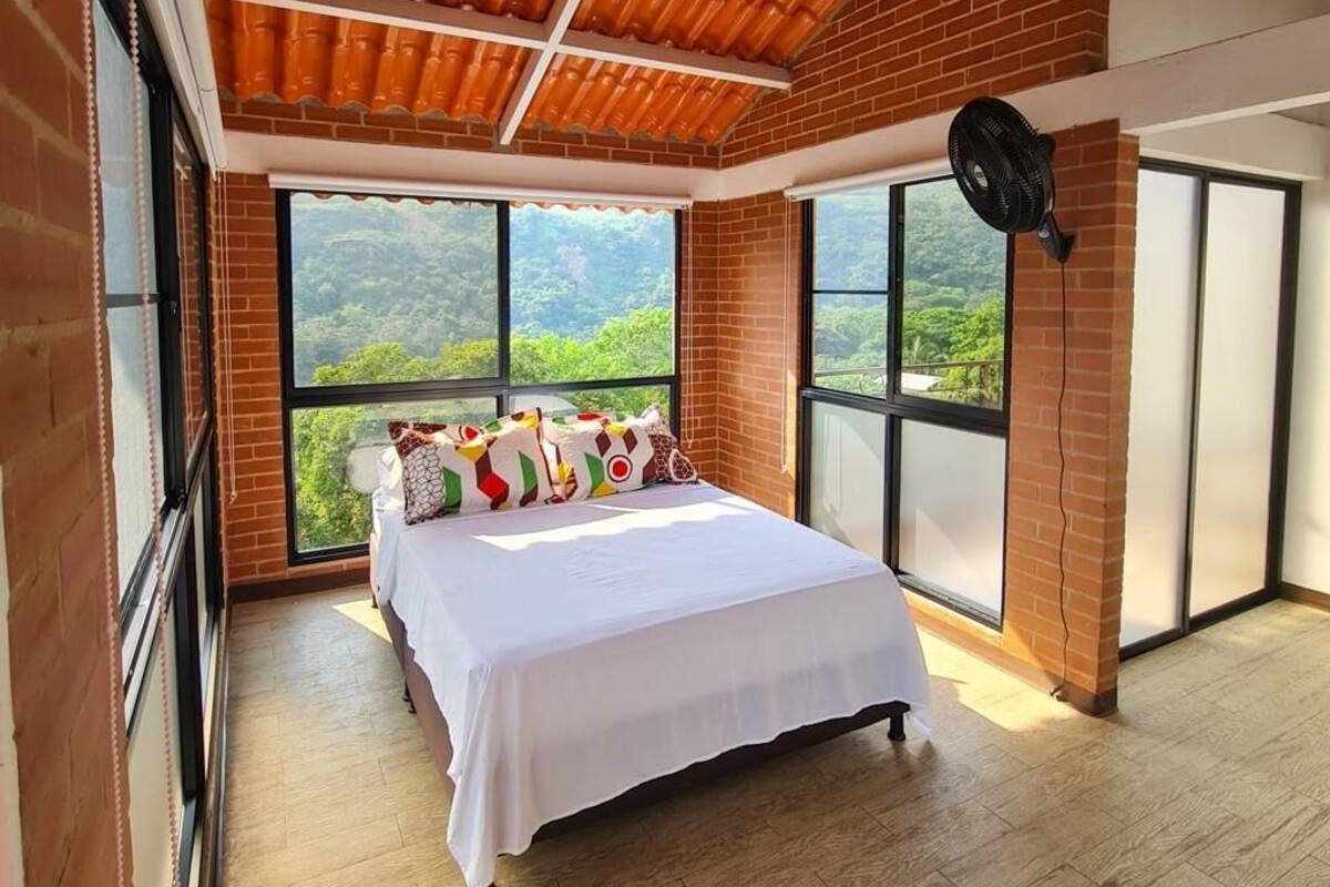 Un dormitorio con una cama grande, un ventilador y vistas a un bosque a través de amplios ventanales.