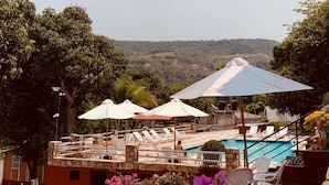 A poolside area with lounge chairs and umbrellas, a stone wall, and a view of a hilly landscape.