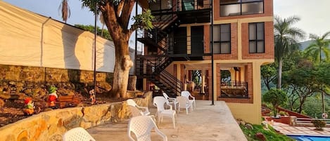 Un edificio de dos pisos con un balcón, zona de estar exterior con sillas de plástico blancas y una piscina al fondo.