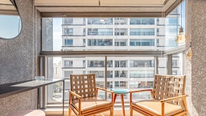 A balcony with wicker chairs and a small table, large windows offering a view of a modern building.