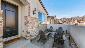 A patio with Adirondack chairs and a table, a stone wall, and a door with a doormat. - Gated PGA West 2023 New Build (EV Charger) (La Quinta)