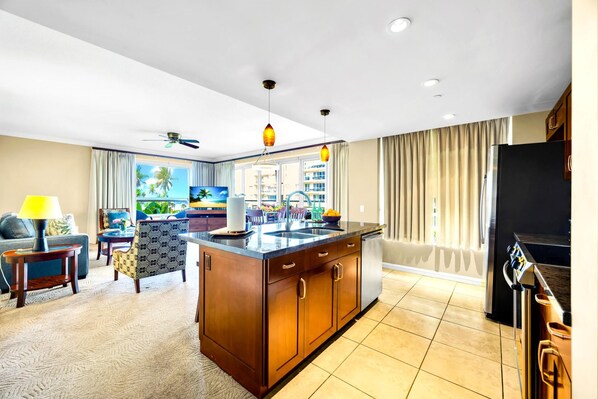 A modern hotel room with a kitchen area, a living area with a sofa and a TV, and a large window with a view of palm trees.